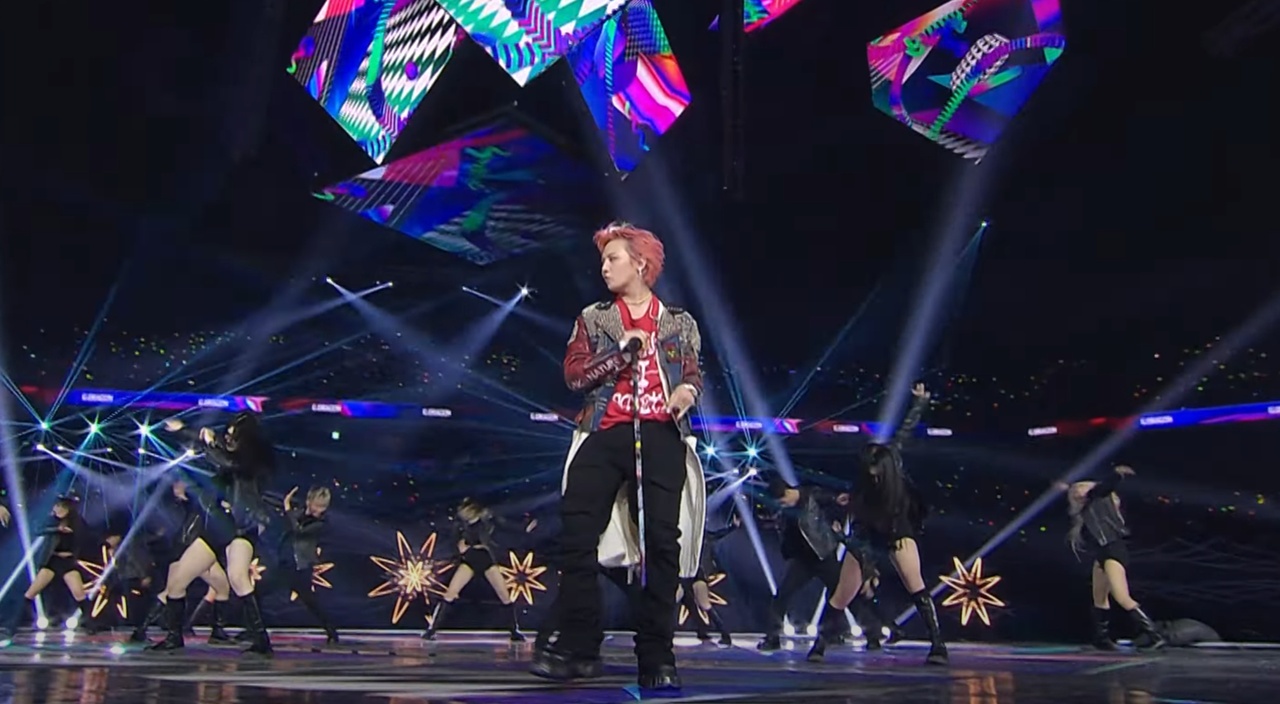K-pop idol performing on a vibrant concert stage with backup dancers, dynamic lighting effects, and a cheering crowd at a large music event.
