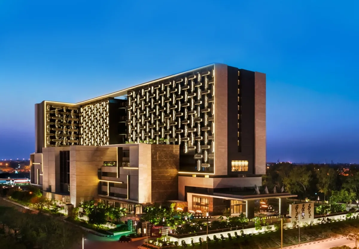 Exterior view of a luxury modern hotel building illuminated at dusk in New Delhi, India, showcasing contemporary architecture and lush landscaping.