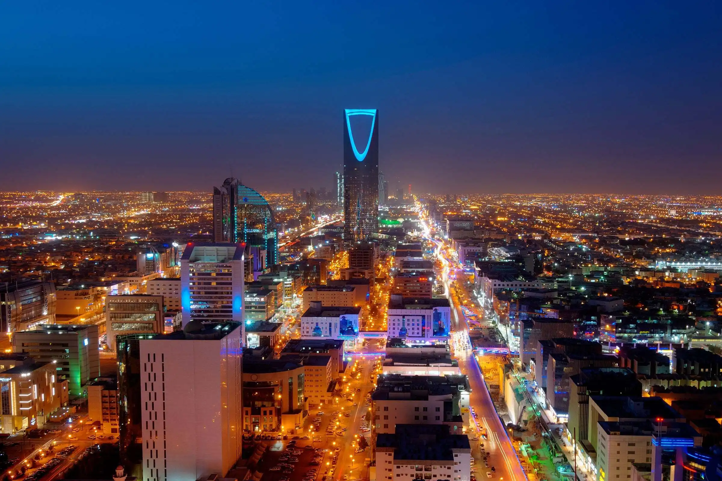Night view of Riyadh cityscape in Saudi Arabia with the iconic Kingdom Centre Tower illuminated, surrounded by bright urban lights and modern architecture.