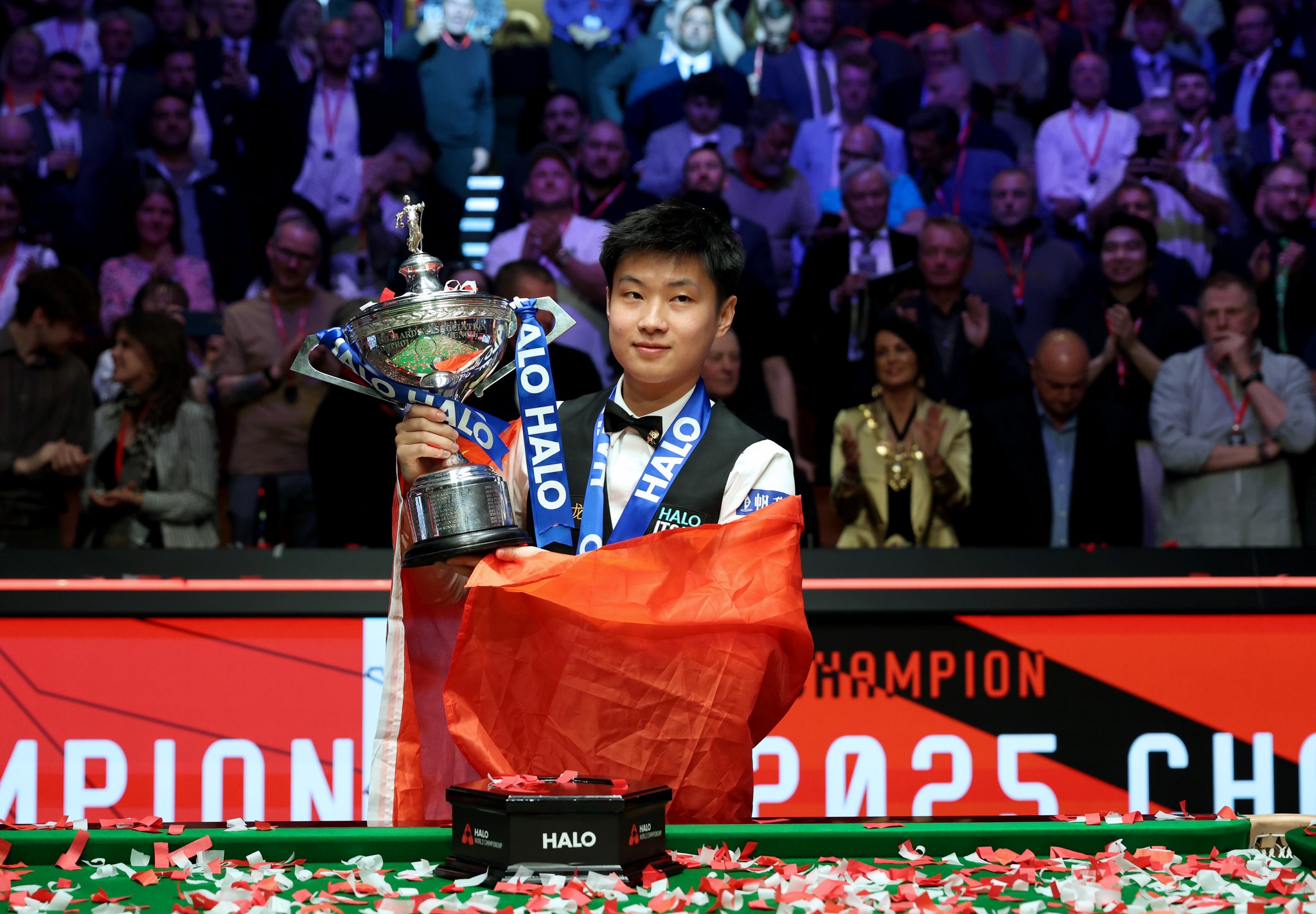 Chinese snooker player holding the 2025 World Championship trophy wrapped in national flag, celebrating victory in front of cheering audience and confetti.