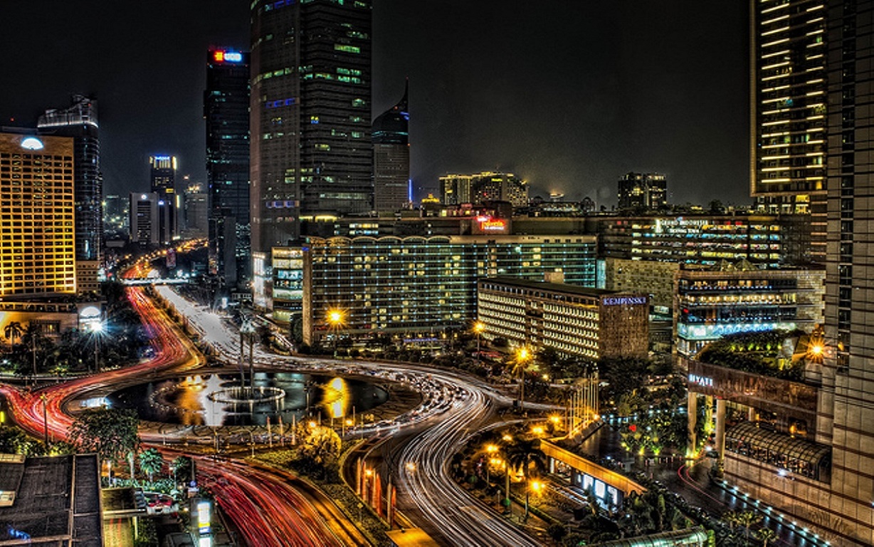 Night view of Jakarta’s cityscape featuring the iconic Hotel Indonesia Roundabout, illuminated skyscrapers, and light trails from busy traffic.