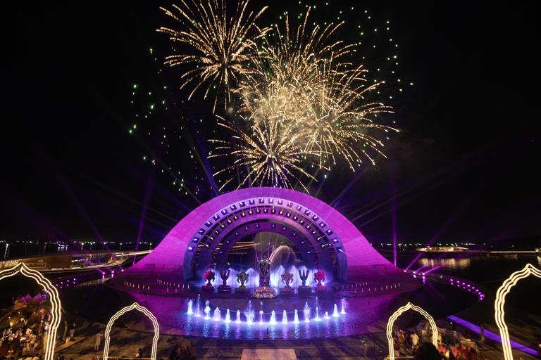 Grand night-time cultural event with vibrant fireworks above a lit-up stage featuring a semi-circular architectural design, fountain display, and traditional dance performances.