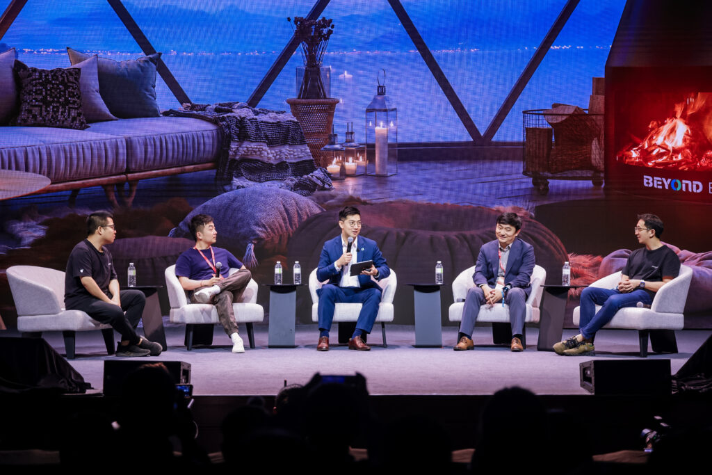 Panel discussion at BEYOND Expo featuring five male speakers in casual and formal attire on a modern stage with cozy lounge decor and digital fireplace backdrop.