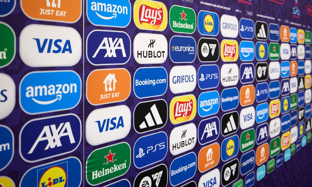 Wall of sponsor logos at a UEFA Women’s EURO 2025 event featuring brands like Amazon, Visa, Lay’s, Booking.com, Adidas, AXA, Heineken, and PlayStation.