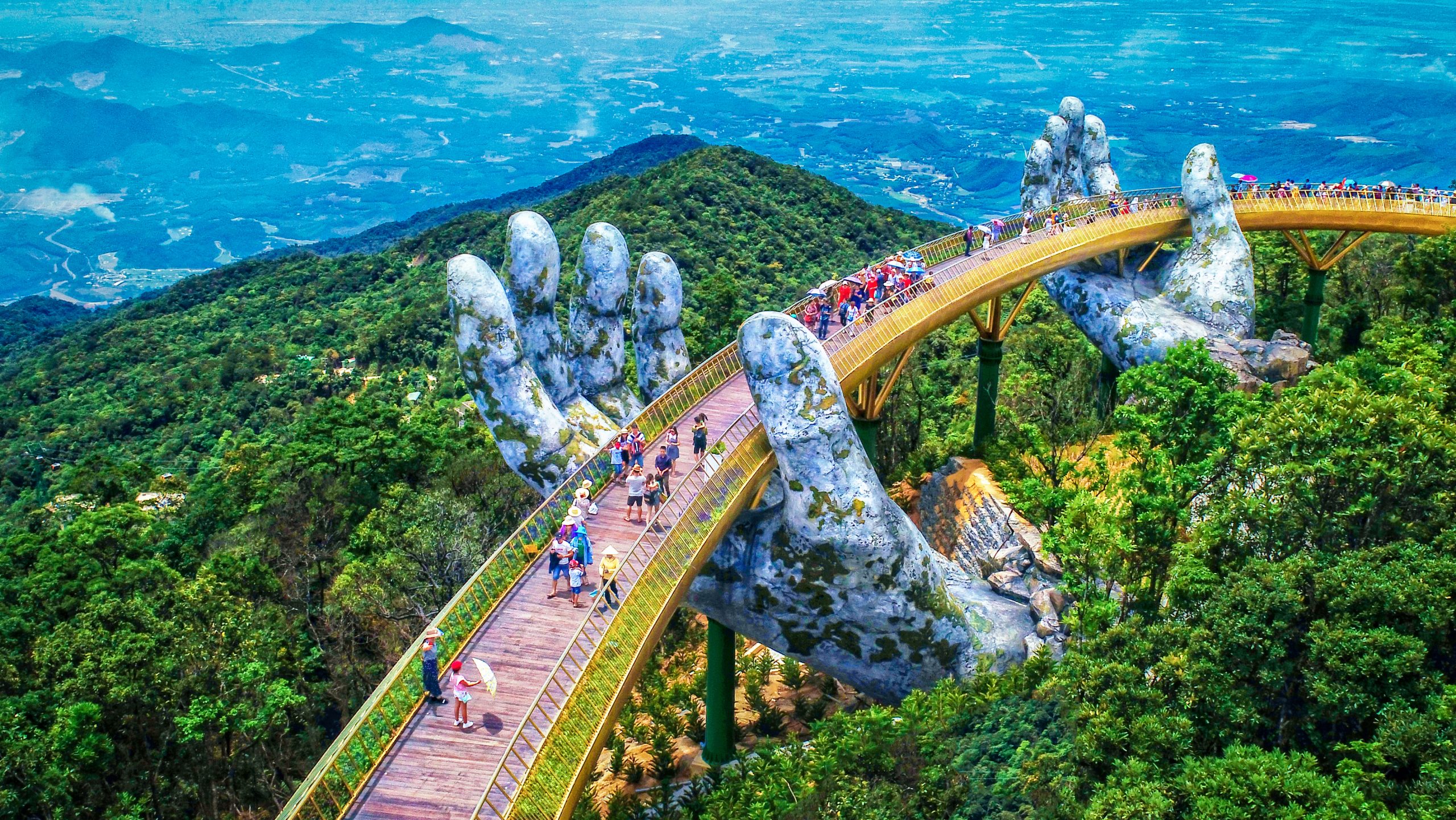 Tourists walking along the Golden Bridge in Da Nang, Vietnam, a scenic mountain walkway held by giant stone hands above lush green forest.