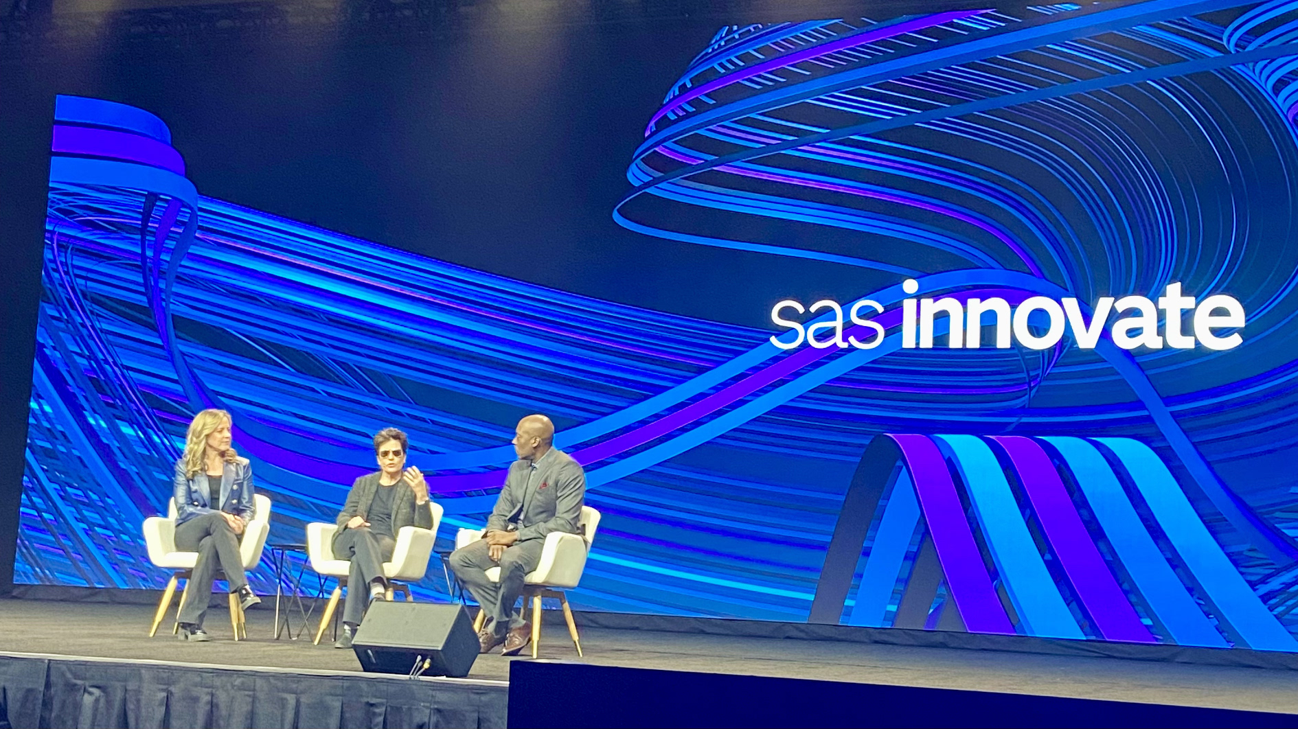 Panel discussion on stage at SAS Innovate conference featuring three speakers seated in front of a dynamic blue digital backdrop with event branding.