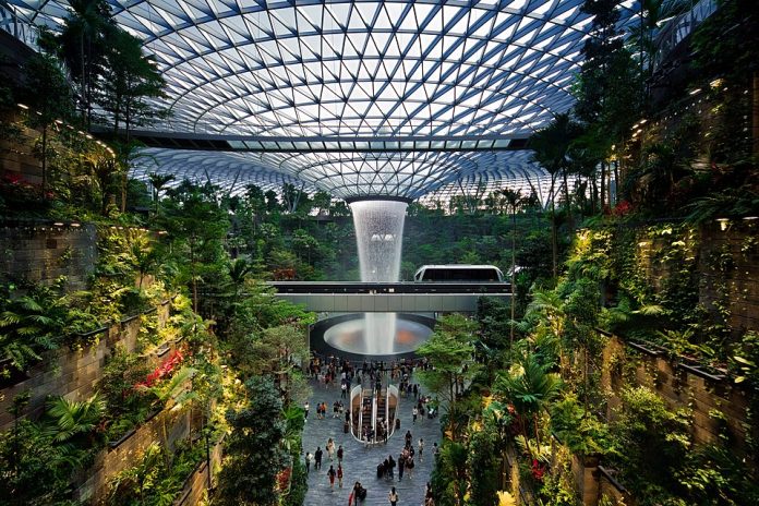 Indoor rainforest and HSBC Rain Vortex waterfall at Jewel Changi Airport in Singapore, surrounded by lush greenery and visitors under a glass dome.