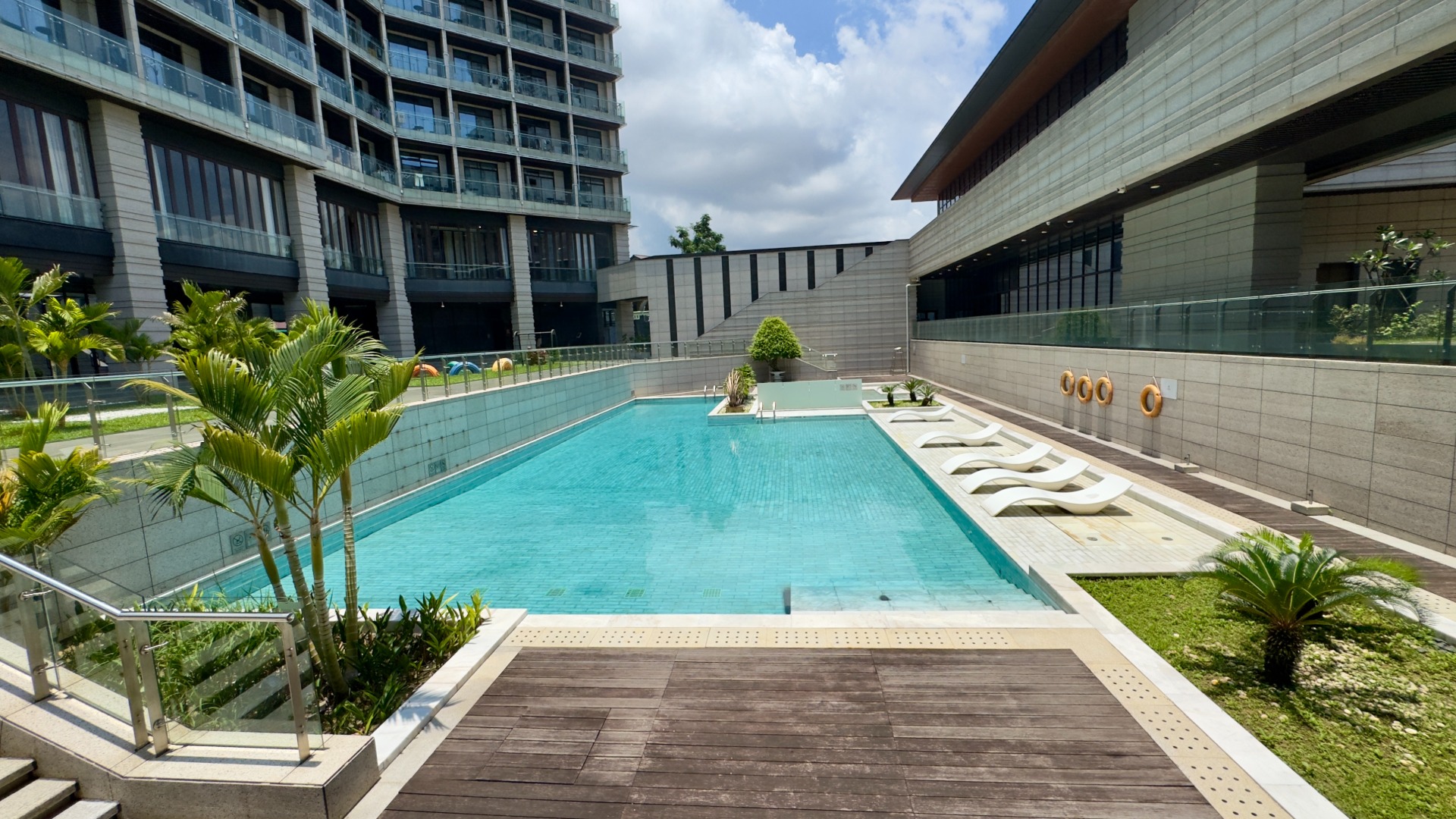 Modern outdoor swimming pool surrounded by luxury apartments and lounge chairs, featuring clean architecture, palm trees, and a bright sunny sky.