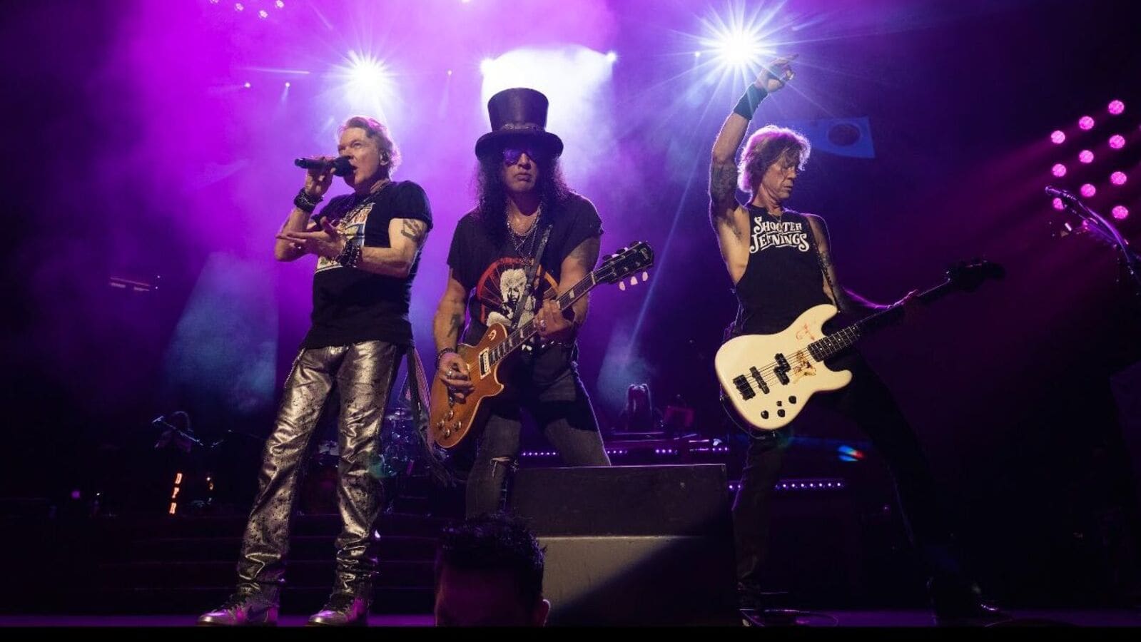 Rock band performing live on stage under dramatic purple lighting, featuring lead singer with microphone, guitarist in top hat, and bassist in concert stance.