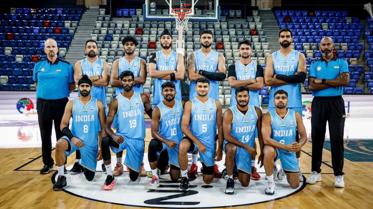 India men’s national basketball team poses for a group photo at the FIBA tournament, wearing light blue jerseys on indoor court with hoop in background.
