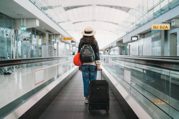 Female traveler with a suitcase and backpack walking through a modern airport terminal on a travelator toward boarding gates.