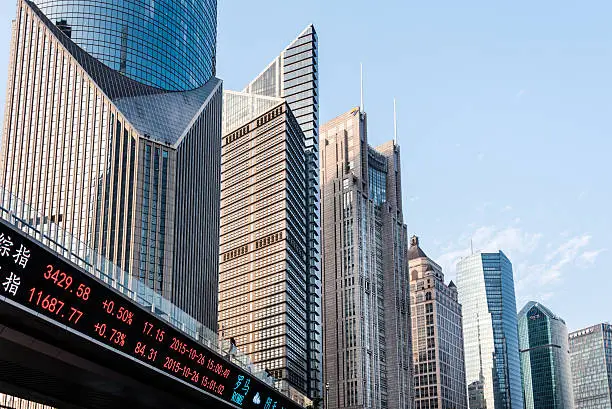 Modern skyscrapers in Shanghai’s financial district with a digital stock ticker display showing market data under a clear blue sky.