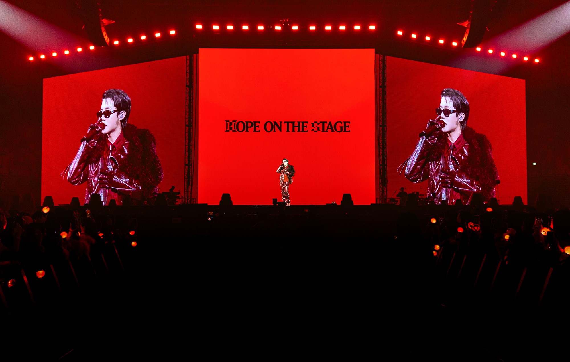 K-pop artist performing on stage in a red outfit with large LED screens displaying “Hope on the Stage” and crowd members holding light sticks during a live concert.
