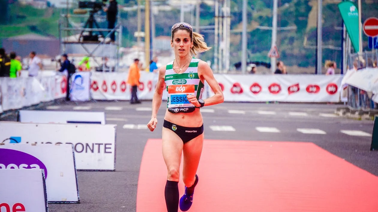 Female athlete running toward the finish line during a road race, wearing a green and white striped sports top and a determined expression on a red track.