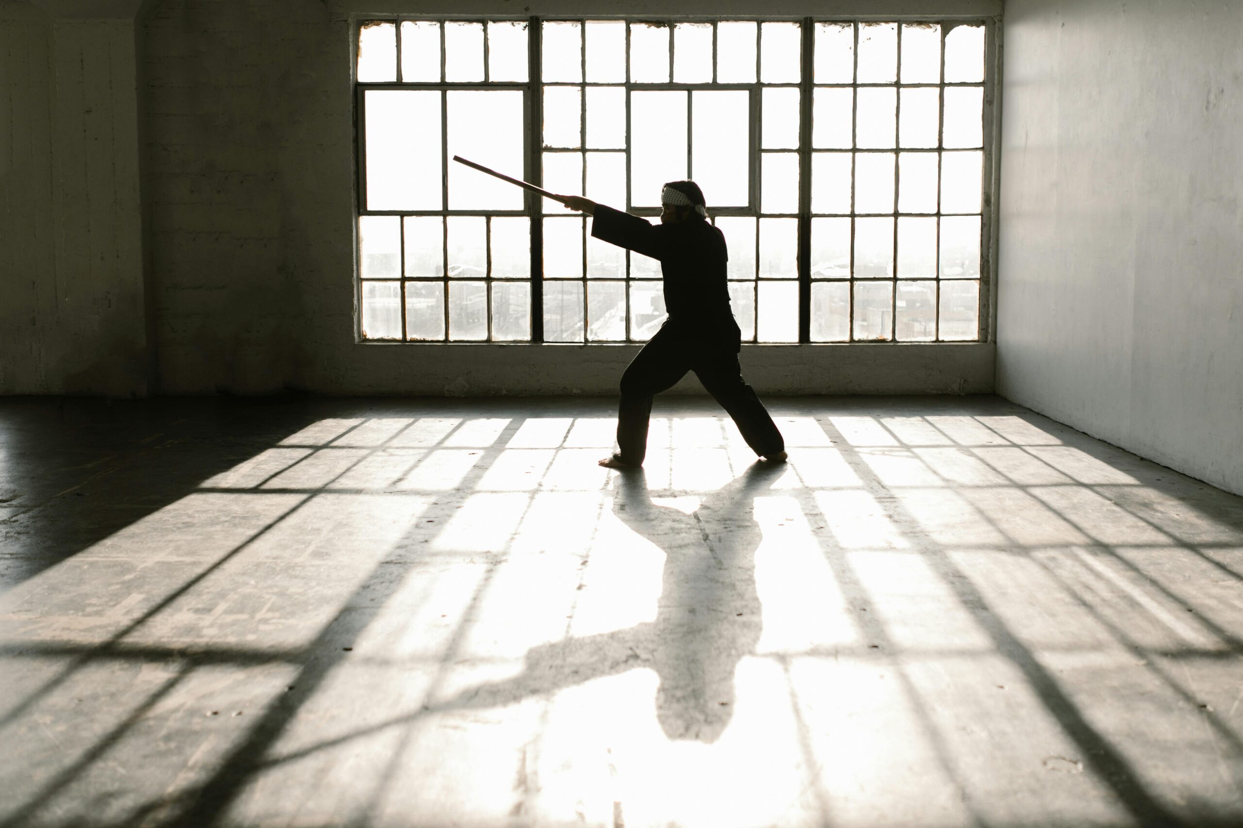 Martial artist practicing with a wooden staff in a sunlit studio, casting a strong shadow on the floor in front of large industrial windows.