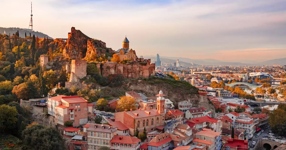 Scenic view of Tbilisi, Georgia featuring the historic Narikala Fortress and colorful Old Town buildings at sunset with a modern city skyline in the background.