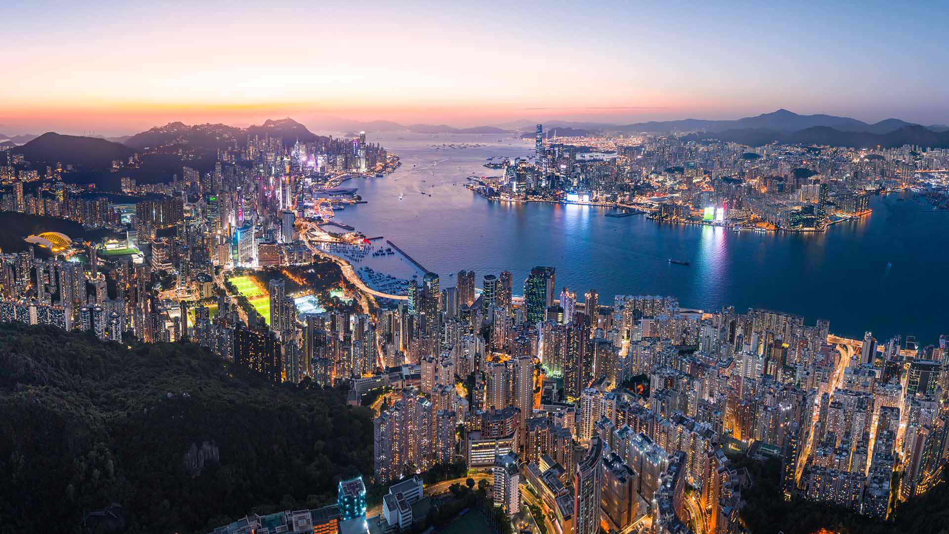 Panoramic aerial view of Hong Kong skyline and Victoria Harbour at dusk with illuminated skyscrapers and mountain backdrop
