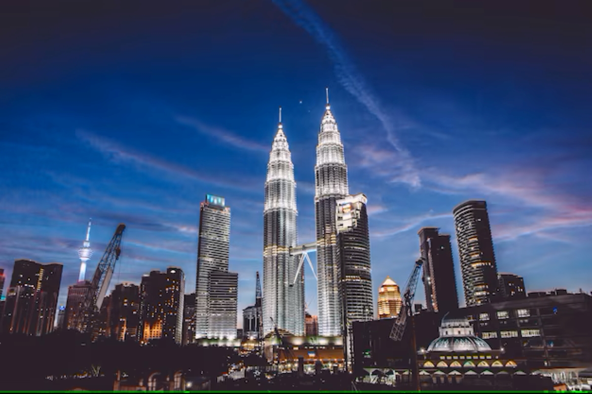 Night skyline of Kuala Lumpur featuring the illuminated Petronas Twin Towers surrounded by modern skyscrapers and construction cranes under a twilight sky in Malaysia.